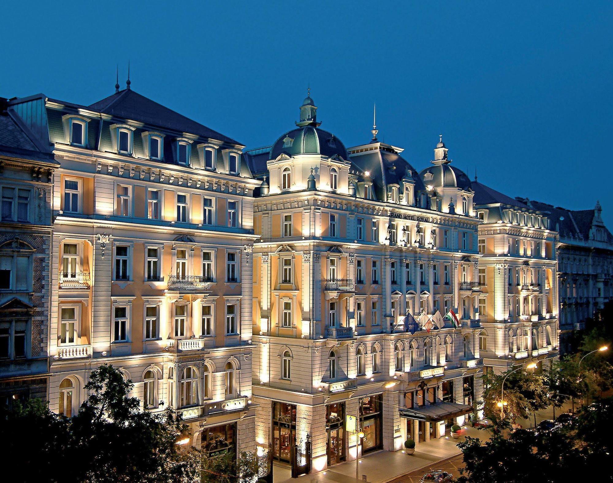 Corinthia Budapest Exterior photo The photo shows a large, elegantly designed building illuminated at dusk. It features architectural elements typical of classic European styles, such as ornate detailing, balconies, and a prominent roofline with domes. The facade is beautifully lit, 